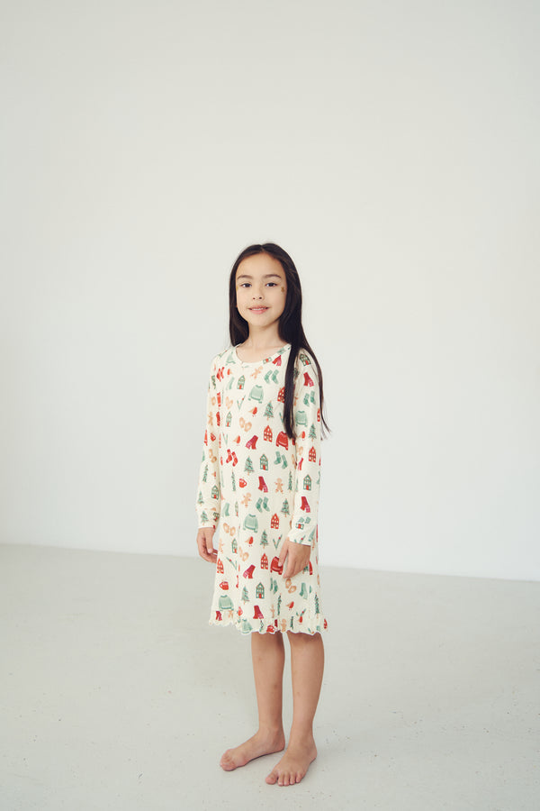 A young girl with long dark hair stands barefoot in a minimalist white studio setting, wearing a long-sleeve cream-colored nightgown decorated with a colorful Christmas tree pattern.