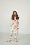 A young girl with long brown hair stands barefoot in a studio wearing a matching long-sleeve pajama set with an allover Christmas tree and snowman print on an off-white background.