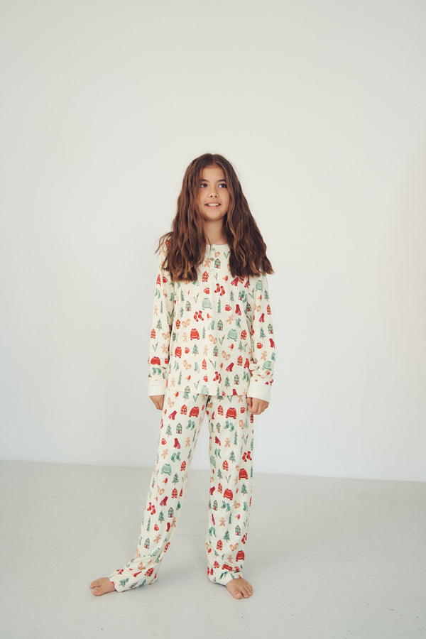 A young girl with long brown hair stands barefoot in a studio wearing a matching long-sleeve pajama set with an allover Christmas tree and snowman print on an off-white background.