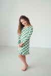 A young girl with long curly brown hair stands barefoot in a bright, minimalist studio setting, wearing a green and white checkered long-sleeve nightgown.