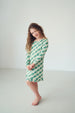 A young girl with long curly brown hair stands barefoot in a bright, minimalist studio setting, wearing a green and white checkered long-sleeve nightgown.