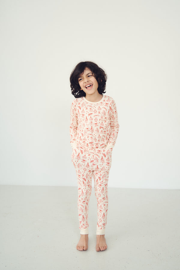 A young child with short dark hair stands barefoot against a plain white studio background, wearing a long-sleeve pajama top and matching pants in a light cream color with a small red and orange allover pattern.