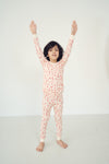 A child wearing a long-sleeve pajama set with an allover red and orange pattern on a cream background, standing with arms raised against a plain white studio background.