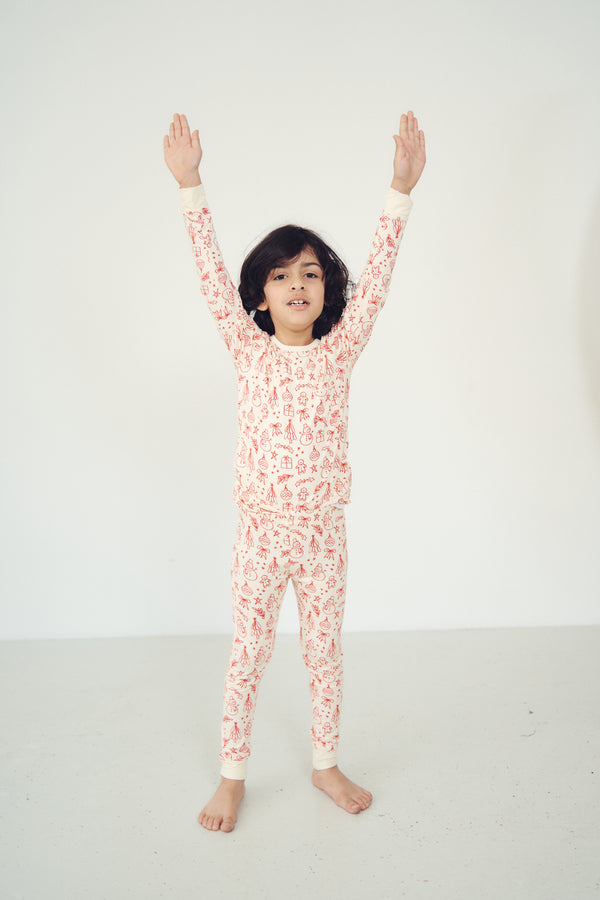 A child wearing a long-sleeve pajama set with an allover red and orange pattern on a cream background, standing with arms raised against a plain white studio background.