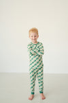 A young boy with blonde hair stands barefoot against a plain white wall, wearing a long-sleeve one-piece pajama set with a green and cream checkerboard pattern.