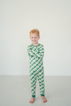 A young boy with red hair stands barefoot against a plain white wall, wearing a long-sleeve pajama set with a green and white checkerboard pattern.