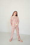 A young girl stands barefoot against a plain white studio wall wearing a long-sleeve plaid pajama top and matching pajama pants in a red, beige, and white pattern.