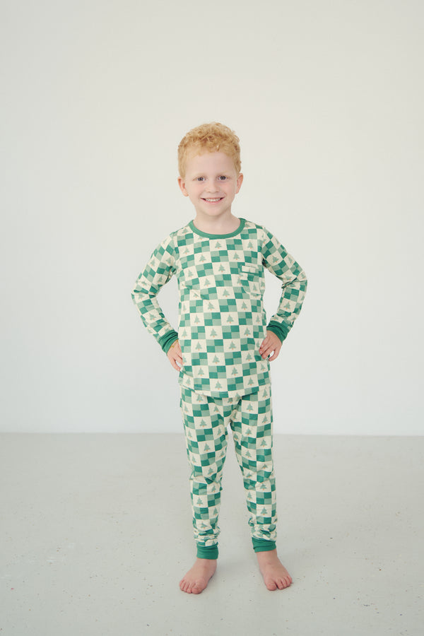 A young boy with red hair stands barefoot in a studio setting, wearing a long-sleeve one-piece pajama set with a green and cream checkerboard pattern.