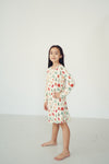 A young girl with long dark hair stands barefoot in a studio, wearing a cream-colored long-sleeve dress with a colorful pattern of red and green illustrations.