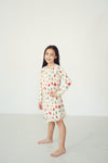 A young girl with long dark hair stands barefoot in a studio setting, wearing a long-sleeve cream-colored nightgown with a festive red, green, and blue holiday print.