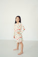 A young girl with long dark hair stands barefoot in a studio setting, wearing a long-sleeve cream-colored nightgown with a festive red, green, and blue holiday print.