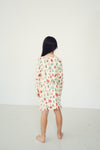 A child standing with their back to the camera, wearing a cream-colored long-sleeve pajama dress with a colorful Christmas tree pattern, set against a plain white studio background.