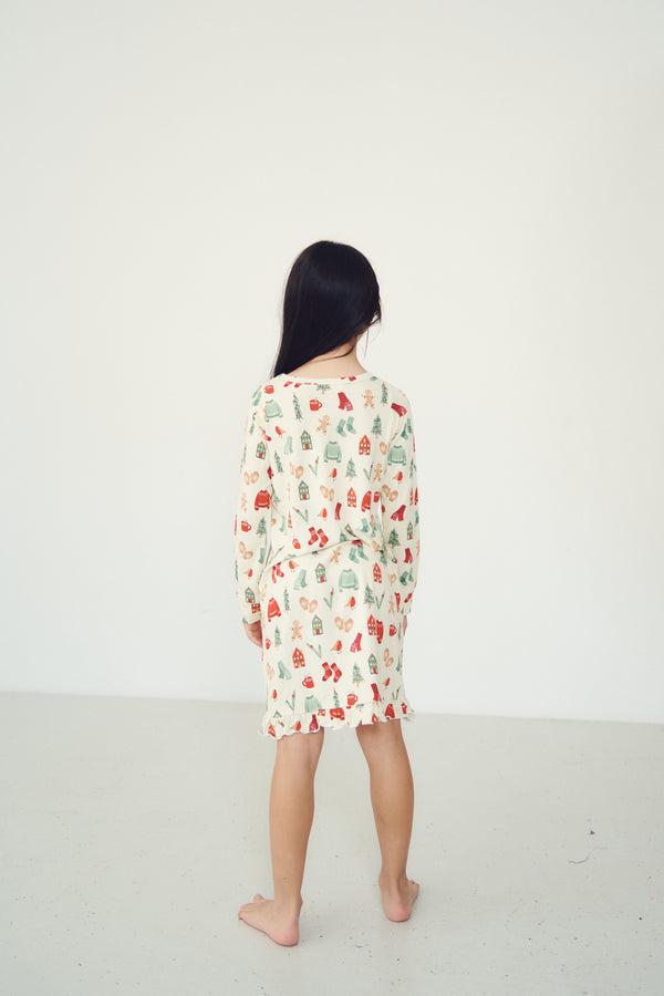A child standing with their back to the camera, wearing a cream-colored long-sleeve pajama dress with a colorful Christmas tree pattern, set against a plain white studio background.