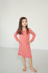 A young girl with long brown hair stands barefoot in a studio, wearing a long-sleeve red and white wavy patterned dress with a ruffled hem.