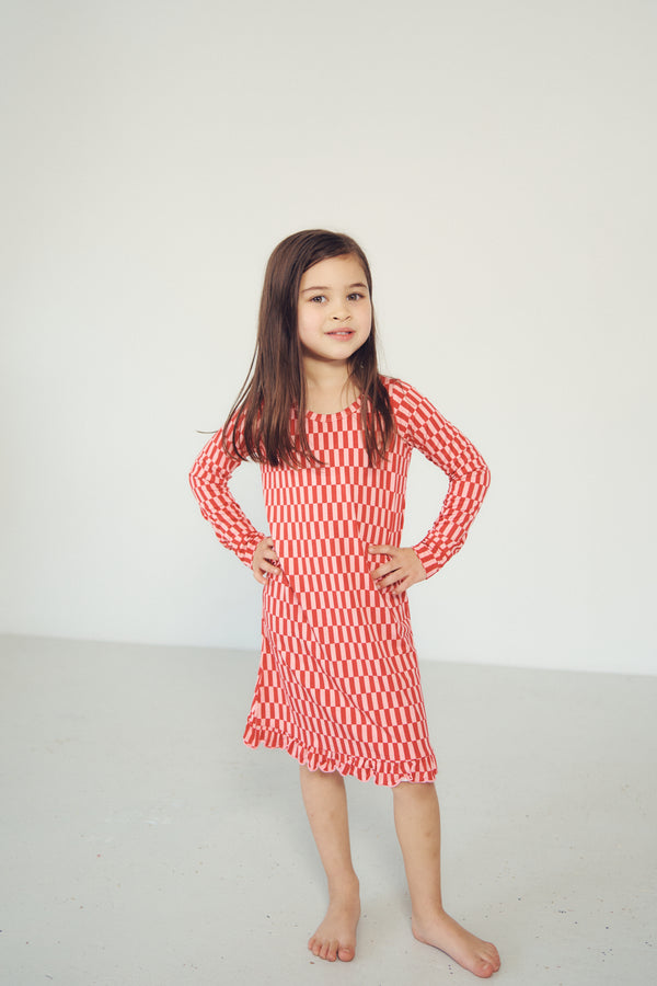 A young girl with long brown hair stands barefoot in a studio, wearing a long-sleeve red and white wavy patterned dress with a ruffled hem.