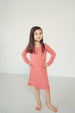 A young girl with long brown hair stands barefoot in a studio, wearing a long-sleeve red and white wavy patterned dress with a ruffled hem.