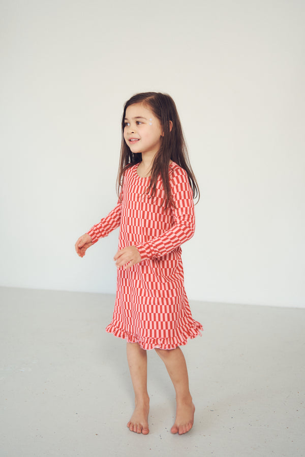 A young girl with long dark hair stands barefoot in a bright studio, wearing a long-sleeve red and white vertically striped dress with a ruffled hem.