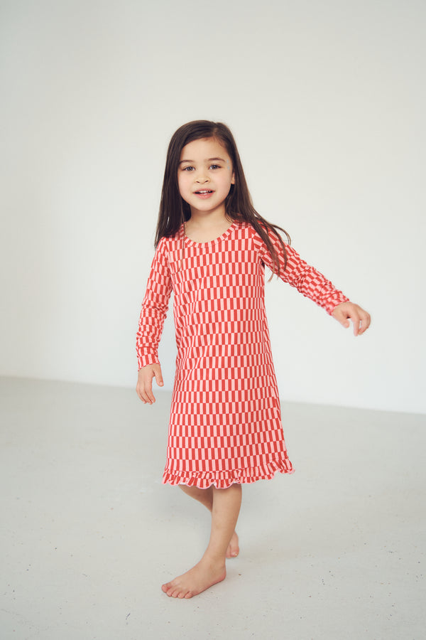 A young girl with long dark hair stands barefoot in a bright studio, wearing a long-sleeve dress with a red and white checkerboard pattern.