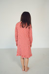 A woman viewed from behind wearing a long-sleeve dress with a red and white checkerboard pattern, standing barefoot against a plain white studio background.
