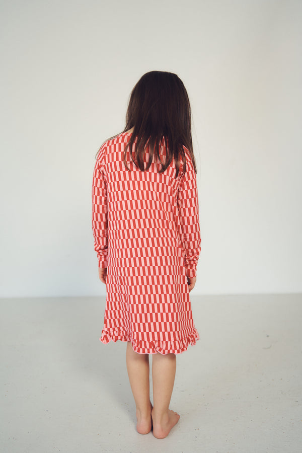 A woman viewed from behind wearing a long-sleeve dress with a red and white checkerboard pattern, standing barefoot against a plain white studio background.