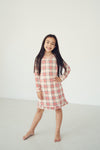 A young girl with long dark hair stands barefoot in a bright white studio, wearing a long-sleeve plaid dress with red, black, and white stripes.