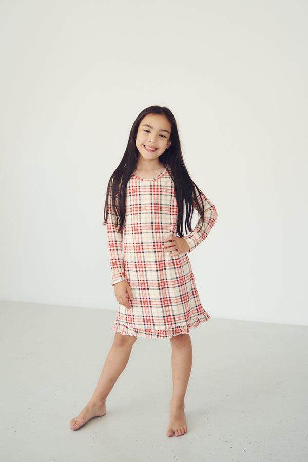 A young girl with long dark hair stands barefoot in a bright white studio, wearing a long-sleeve plaid dress with red, black, and white stripes.