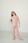 A young girl stands barefoot in a studio wearing a long-sleeve plaid pajama top and matching pajama pants in a red, white, and light blue check pattern.