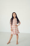 A young girl with long dark hair stands barefoot in a bright, minimalist studio setting, wearing a long-sleeve plaid dress with red, white, and beige stripes.