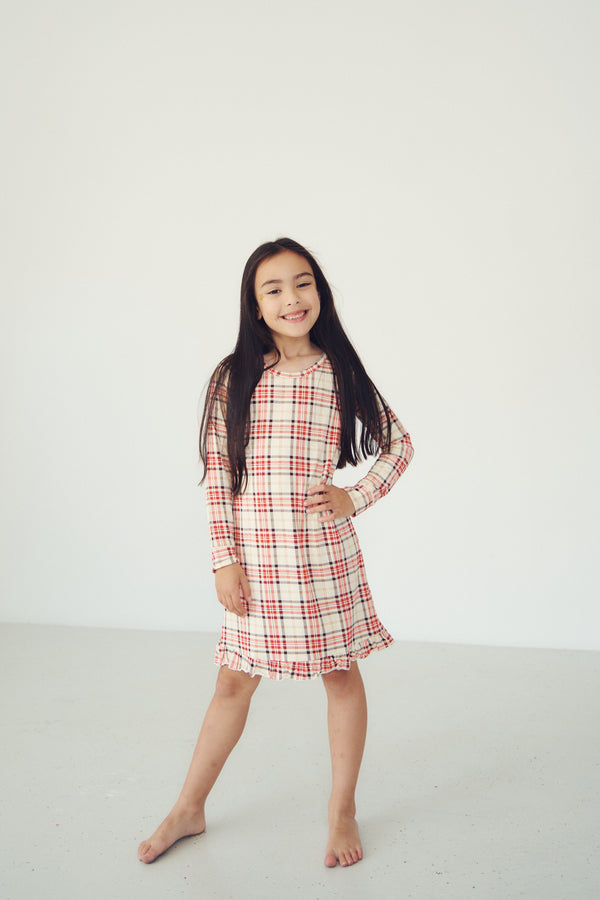 A young girl with long dark hair stands barefoot in a bright, minimalist studio setting, wearing a long-sleeve plaid dress with red, white, and beige stripes.