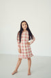 A young girl with long dark hair stands barefoot in a bright, minimalist studio setting, wearing a long-sleeve plaid dress with red, white, and beige stripes.