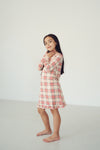 A young girl with long dark hair stands barefoot in a minimalist studio setting, wearing a long-sleeve plaid dress with a cream base and red and orange stripes.
