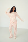 A woman modeling a long-sleeve, one-piece pajama jumpsuit with a pink and white abstract pattern, set against a plain white studio background.
