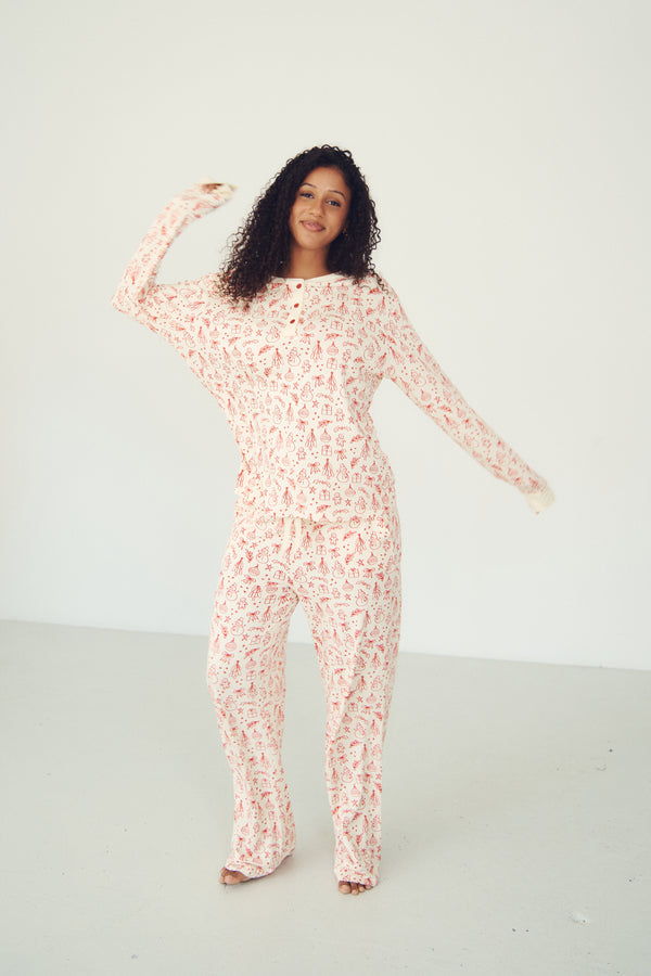 A woman modeling a long-sleeve, one-piece pajama jumpsuit with a pink and white abstract pattern, set against a plain white studio background.