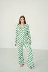 A woman modeling a matching green and white checkered pajama set consisting of a long-sleeve button-up top and wide-leg pants, standing against a plain light gray studio background.