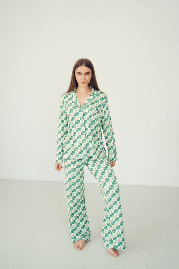 A woman modeling a matching green and white checkered pajama set consisting of a long-sleeve button-up top and wide-leg pants, standing against a plain light gray studio background.