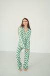 A woman modeling a matching green and white checkered pajama set, consisting of a long-sleeve button-up top and matching pants, standing against a plain white studio background.