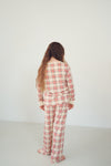 A woman with long light brown hair stands with her back to the camera wearing a matching long-sleeve plaid pajama set in cream, red, and white. The set features a red and white plaid pattern with cream ribbed trim on the collar and cuffs.