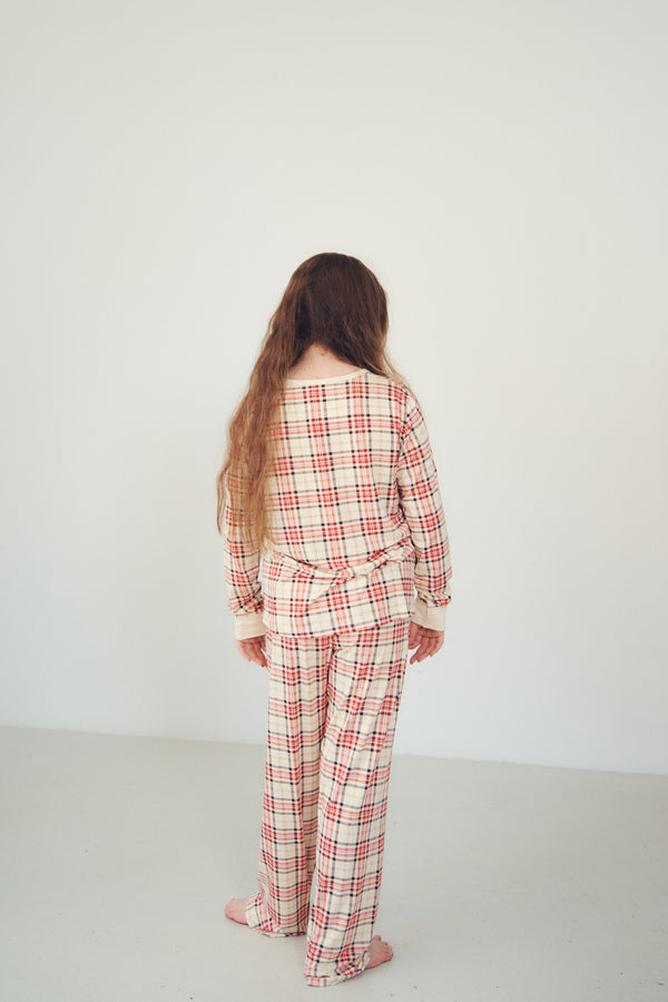 A woman with long light brown hair stands with her back to the camera wearing a matching long-sleeve plaid pajama set in cream, red, and white. The set features a red and white plaid pattern with cream ribbed trim on the collar and cuffs.