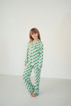 A young girl stands barefoot in a bright white studio setting, wearing a green and white checkered long-sleeve pajama set.