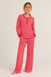 A young girl stands barefoot in a studio wearing a long-sleeve plaid pajama top and matching pajama pants in a red abstract pattern.