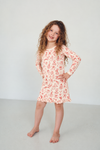 A young girl with long curly brown hair stands barefoot in a bright, minimalist studio setting, wearing a red and white holiday print long-sleeve nightgown.