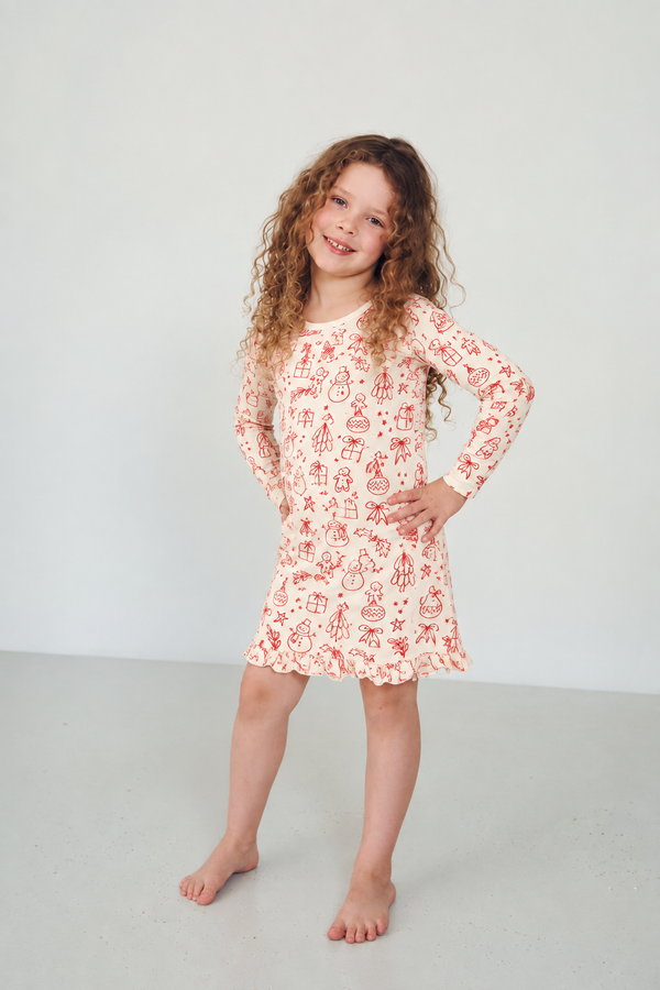 A young girl with long curly brown hair stands barefoot in a bright, minimalist studio setting, wearing a red and white holiday print long-sleeve nightgown.