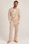 A man wearing a holiday/winter patterned long-sleeve pajama top and matching pants, standing barefoot against a plain light gray studio background.